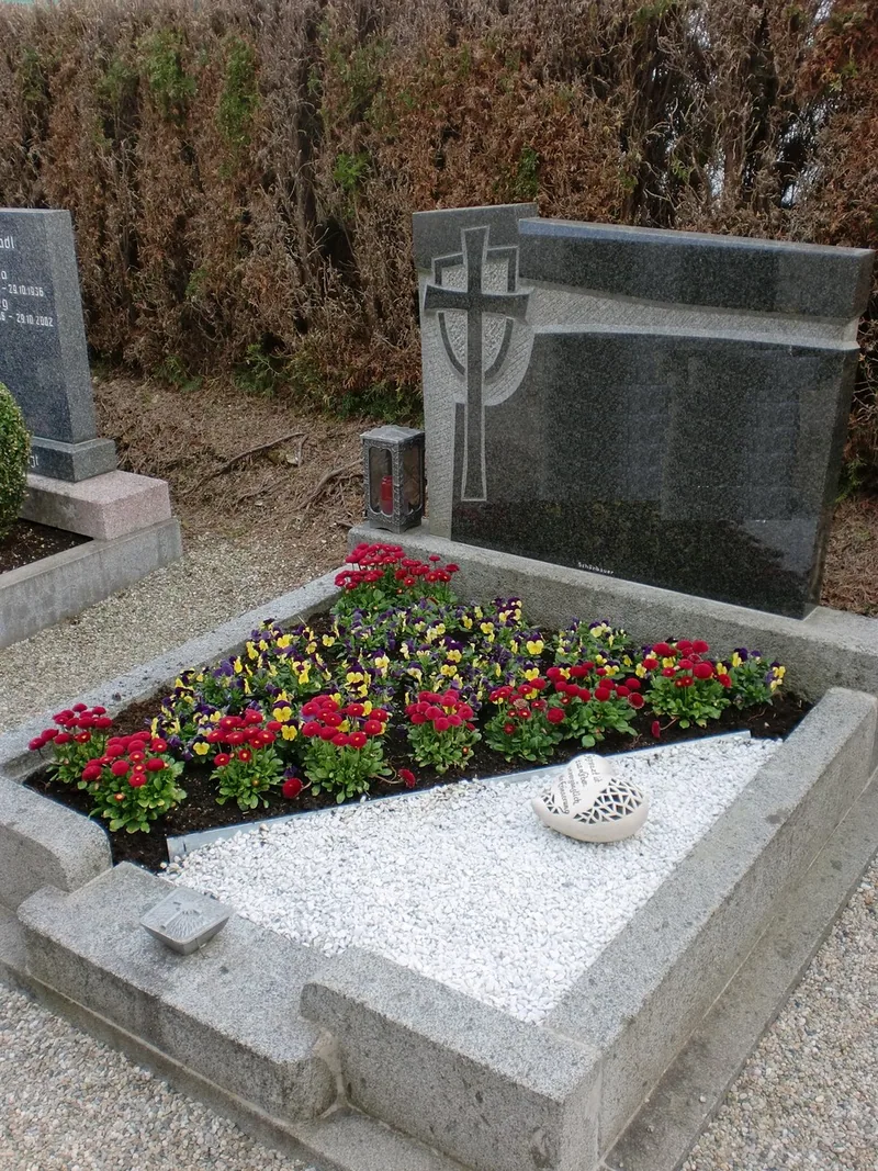 Grab mit roten Begonien und weißen Blüten vor einem dunklen Grabstein